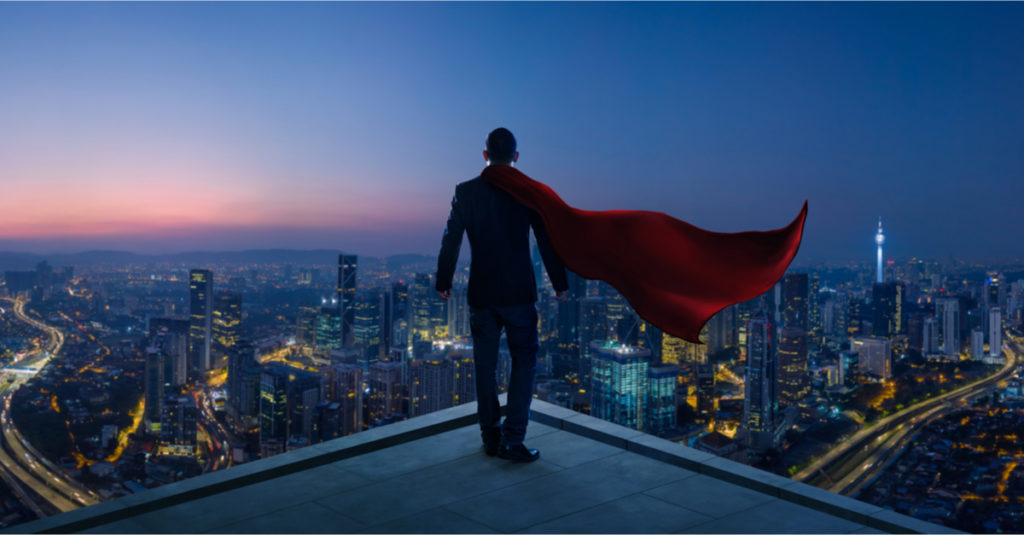 A man wearing a black suit and a red superhero cape stares out at a large city from a atop a tall building.